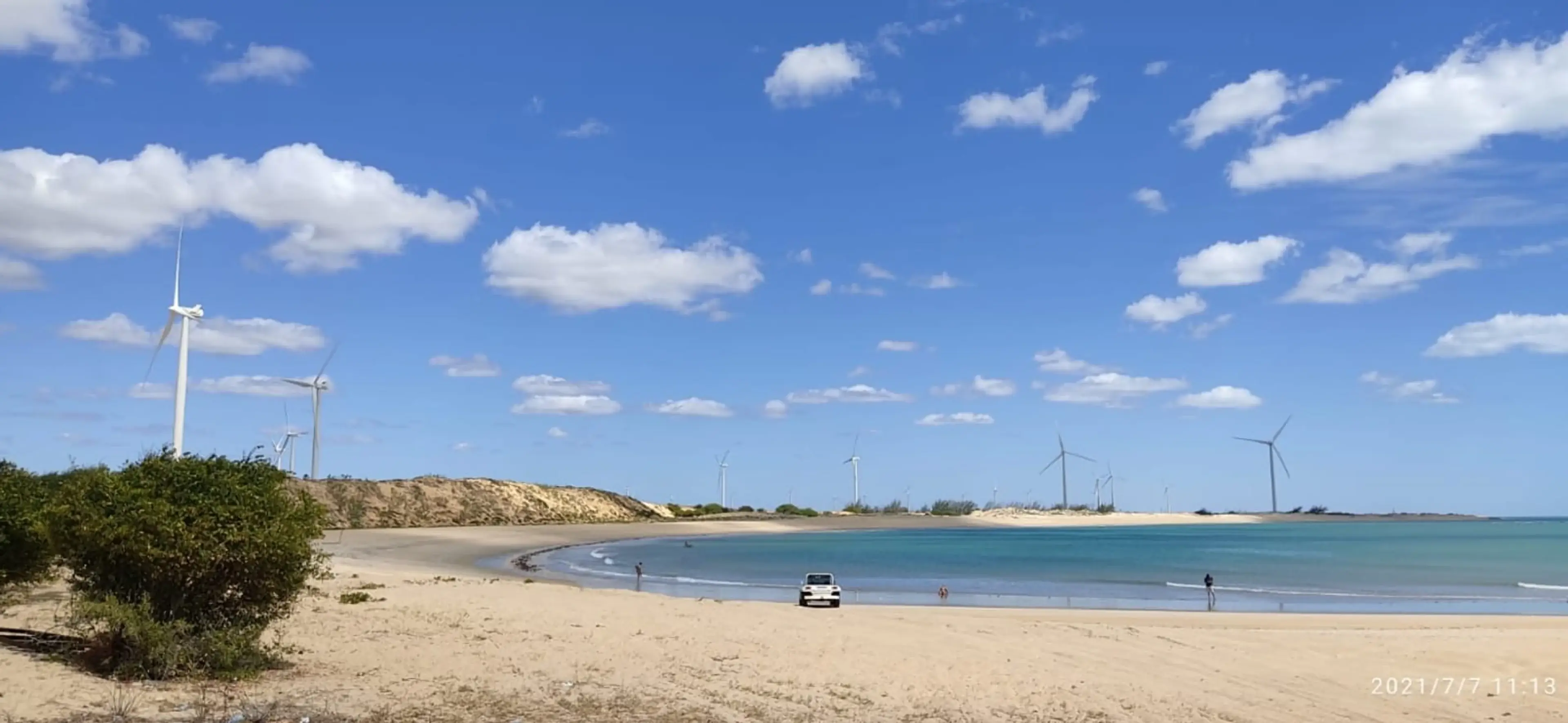 Praia ampla com dunas, mar azul e aerogeradores ao fundo